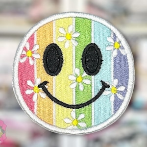 May include: Embroidered patch with a smiley face design. The face has a rainbow striped background, black eyes, and a curved black mouth. White daisy flowers are scattered around the face. The patch is round with a white border. Text at the bottom reads "HOBBY JOBBY CREATIONS".