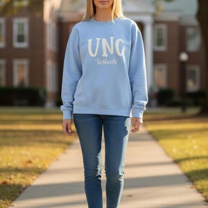 Op de afbeelding: Een lichtblauw sweatshirt met de tekst "UNC Tarheels" in het wit. De persoon draagt een blauwe spijkerbroek en witte sneakers. De achtergrond omvat een bakstenen gebouw en een met bomen omzoomde loopbrug.