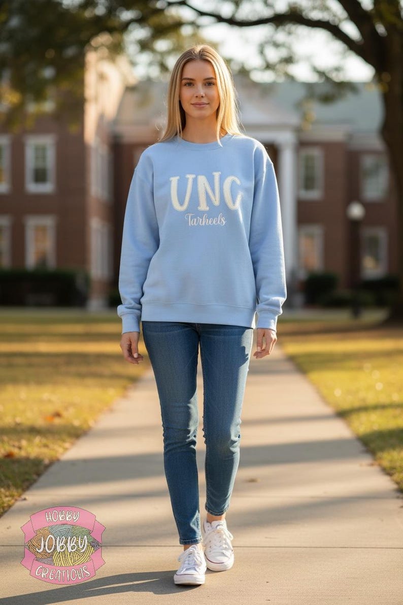 Op de afbeelding: Een lichtblauw sweatshirt met de tekst "UNC Tarheels" in het wit. De persoon draagt een blauwe spijkerbroek en witte sneakers. De achtergrond omvat een bakstenen gebouw en een met bomen omzoomde loopbrug.