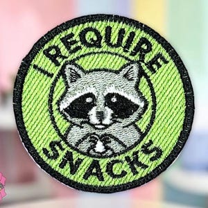 May include: Embroidered patch with a green background and a black border. The patch features a raccoon illustration with the text "I REQUIRE SNACKS" in a circular design. The patch is round.