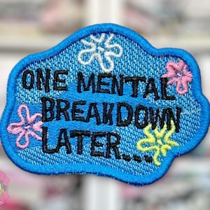 May include: Embroidered patch with the text "ONE MENTAL BREAKDOWN LATER..." in black on a blue denim-look background. The patch has a blue border and is decorated with embroidered white, pink, and yellow floral designs.