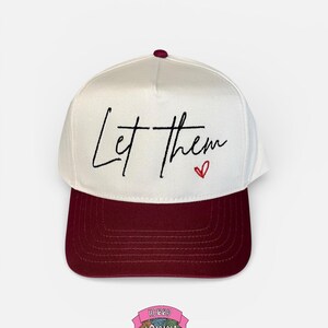 May include: A white baseball cap with a maroon brim and top button. The cap features the handwritten text "Let them" with a small red heart. The cap is a stylish accessory.