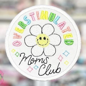 May include: White embroidered patch with the words "OVERSTIMULATED" in rainbow colors around a daisy with a yellow center and a smiling face. The words "Moms Club" are written in black script below the flower.