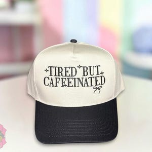 May include: A white and black trucker hat with the text "TIRED BUT CAFFEINATED" in black script. The hat has a black brim and a white front panel. A small bow is below the text. The hat is a casual accessory.
