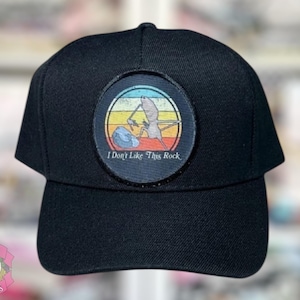 May include: Black baseball cap with a circular patch showing a cartoon figure with a pickaxe, a rock, and the text "I Don't Like This Rock" against a sunset background.