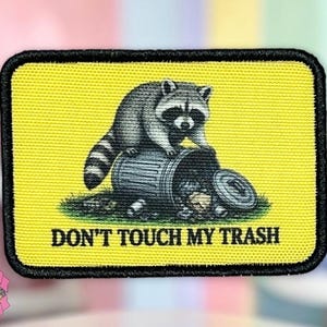 May include: Embroidered patch with a raccoon perched atop a tipped-over trash can. The patch has a yellow background with the text "DON'T TOUCH MY TRASH" in black. The patch is rectangular with a black border.