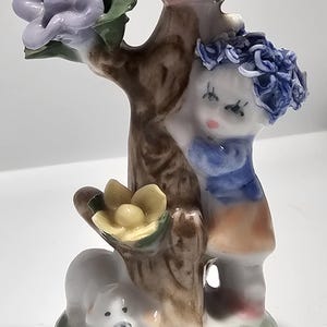 May include: A ceramic figurine depicting a girl with a blue floral crown, standing next to a tree with pink, purple, and yellow flowers. A small white animal rests at the base of the tree on a green grassy base. The figurine is hand-painted.