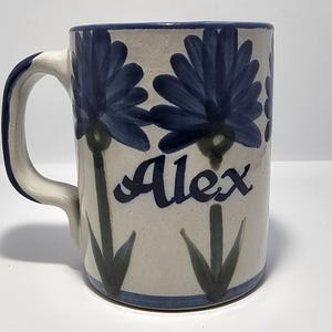Puede incluir: Una taza de cerámica con base blanca, adornada con diseños florales azules y el nombre "Alex" en escritura azul. La taza presenta un borde, asa y detalles azules, creando un aspecto cohesivo y decorativo.