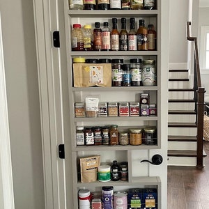 May include: A gray, built-in spice rack installed on a door, filled with various jars, bottles, and containers of spices, sauces, and condiments. The rack has multiple shelves and a black door handle. The background shows a staircase and hardwood flooring.
