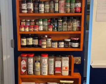 May include: A wooden spice rack with multiple shelves filled with various jars and bottles of spices, herbs, and other food items. The rack is mounted on a door and has a blue background.
