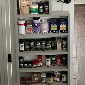 May include: A light gray, multi-tiered wooden spice rack mounted on a door, featuring five horizontal shelves with thin wire guards. The shelves are filled with various spice jars, tea boxes, and food containers of different shapes, sizes, and colors.