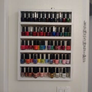 May include: A white wooden shelf unit with five shelves holding rows of colourful nail polish bottles. The bottles are arranged in rows and are mostly in shades of red, blue, green, and purple.