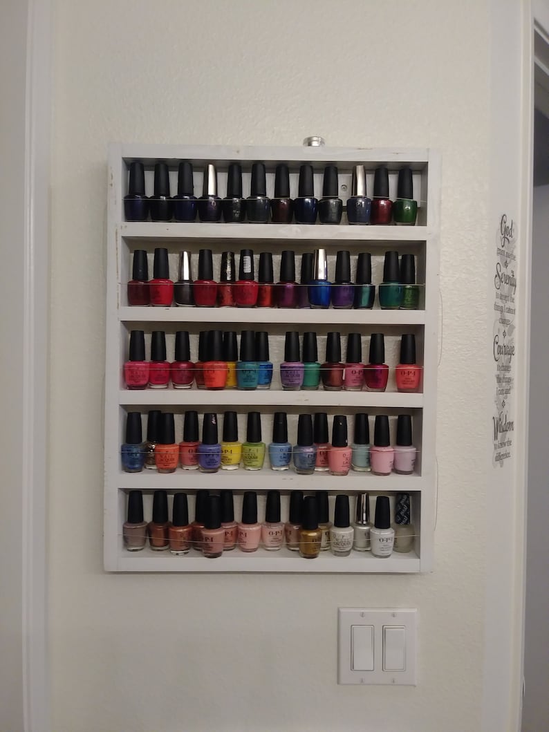 May include: A white wooden shelf unit with five shelves holding rows of colourful nail polish bottles. The bottles are arranged in rows and are mostly in shades of red, blue, green, and purple.
