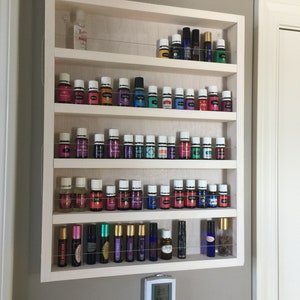 May include: White wooden shelf unit with five shelves, each holding rows of small glass bottles with colourful liquids. The bottles are arranged in a grid pattern. The shelf unit is mounted on a wall.