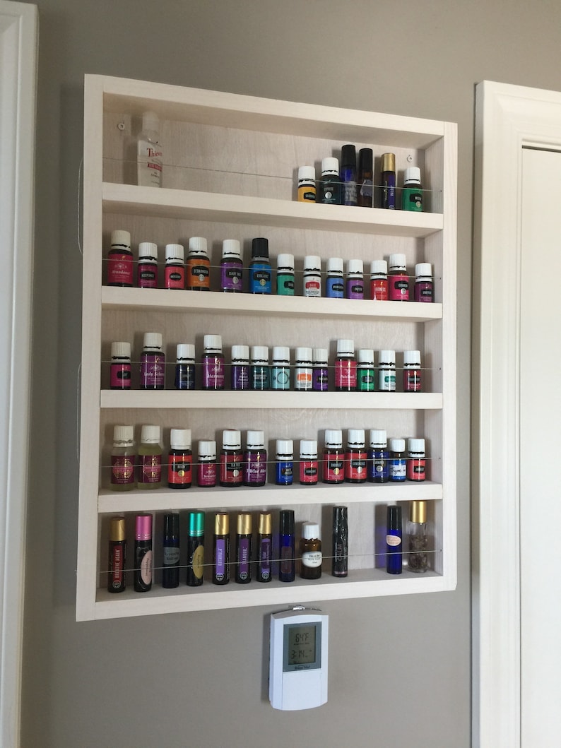 May include: White wooden shelf unit with five shelves, each holding rows of small glass bottles with colourful liquids. The bottles are arranged in a grid pattern. The shelf unit is mounted on a wall.