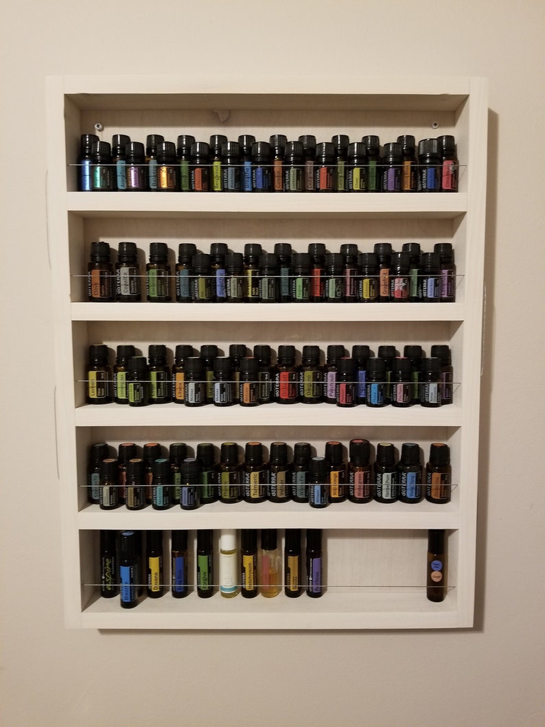 May include: White wooden shelf unit with five shelves, each filled with rows of small glass bottles with black caps. The bottles are filled with colourful liquids.