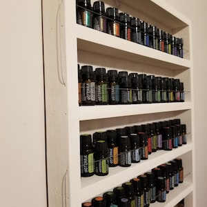 May include: A white wooden shelf unit with five shelves, each filled with rows of small, dark glass bottles with colourful labels. The labels have the word "dōTERRA" on them.