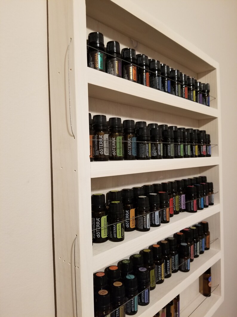 May include: A white wooden shelf unit with five shelves, each filled with rows of small, dark glass bottles with colourful labels. The labels have the word "dōTERRA" on them.