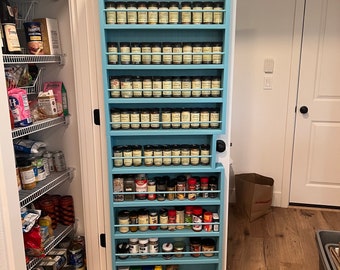 May include: A tall, light blue wooden spice rack mounted on a door, featuring multiple horizontal shelves with protective rails. The rack is filled with numerous spice jars of varying sizes, shapes, and label colours, organised in neat rows.
