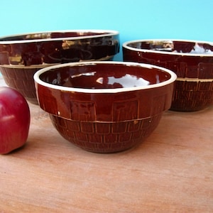 May include: Three brown ceramic bowls with a basket weave design and white rims are displayed. A red apple sits to the left of the bowls. The bowls are of varying sizes and are arranged on a wooden surface.