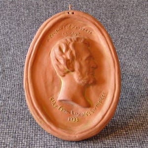 Puede incluir: Una placa ovalada de arcilla roja con un retrato en relieve de Abraham Lincoln. La placa dice "A Century of Progress Chicago 1933 Clay from Lincoln's Birthplace".