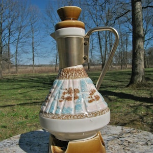 May include: A white ceramic liquor decanter with a gold metal top and a brown base. The decanter has a blue and brown floral pattern and a handle.