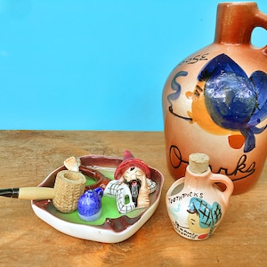 May include: A set of three ceramic figurines, each depicting a whimsical character. The largest figurine is a brown jug with a blue and orange painted design and the text "Goose Creek". The second figurine is a brown and green ashtray with a reclining figure holding a pipe. The third figurine is a small brown jug with a blue and white painted design and the text "Toothpicks".