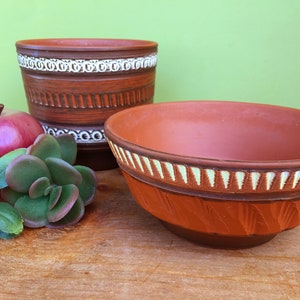 May include: Two terracotta pots with white geometric designs. The larger pot is taller and has a wider opening. The smaller pot is shorter and has a narrower opening. Both pots have a brown glaze.