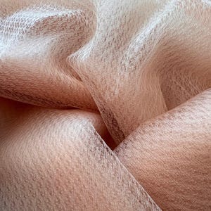 Op de afbeelding: Close-up van een vloeiende, transparante stof in tinten perzik en crème. Het materiaal is delicaat getextureerd, met een subtiele glans en een zachte, luchtige uitstraling. De plooien en lagen creëren diepte en visuele interesse, wat duidt op een luxe textiel.