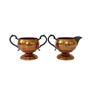 May include: A copper sugar bowl and creamer set. The bowl has two black handles and a pedestal base. The creamer has a spout and a similar design. Both pieces have a shiny, reflective surface.