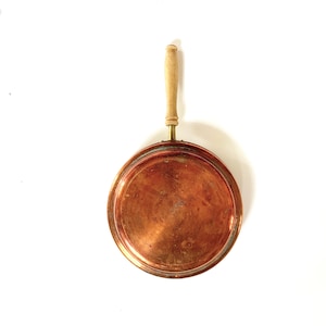 Pode incluir: Uma panela de cobre com uma pega de madeira. A panela é redonda e tem uma superfície brilhante.