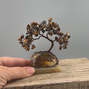 May include: A decorative wire tree sculpture featuring polished tiger's eye gemstone chips as leaves. The twisted brown wire branches emerge from a central stone base, which is mounted on a flat, rectangular, light-colored stone pedestal.
