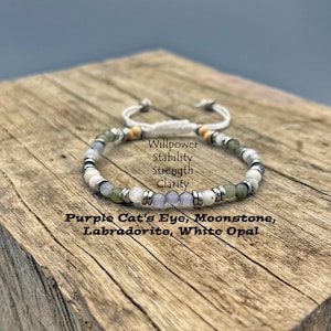 May include: A beaded bracelet featuring purple cat's eye, moonstone, labradorite, and white opal gemstones. The bracelet has a white adjustable cord and silver-colored metal accents. Text on the image reads: Willpower, Stability, Strength, Clarity.