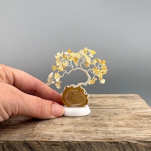 May include: A wire tree sculpture with yellow gemstone leaves and a brown agate base. The tree sits on a small white stone, displayed on a wooden surface. The sculpture is held by a hand.