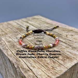 May include: A brown leather cord bracelet with beads of coffee snowflake obsidian, brown jade, cherry quartz, and Australian zebra jasper. The bracelet has a sliding knot closure. The text "Protection, Comfort, Hope, Security" is written on a white background.