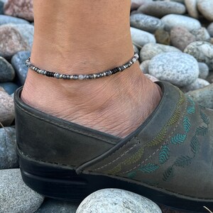 May include: A silver, black, and gray beaded anklet with a delicate design. The anklet is worn on a person's ankle.