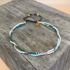 May include: A delicate double-strand anklet with white, light blue, and green beads. The anklet features a gold charm in the shape of a four-leaf clover.