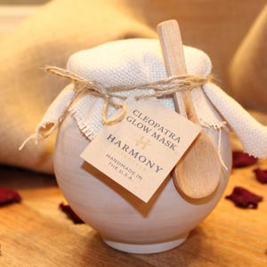 May include: A small, off-white ceramic jar with a fabric lid tied with twine, holding a skincare product. A wooden spoon rests on the jar. A tag reads "Cleopatra Glow Mask" and "Harmony, Handmade in the U.S.A." Dried red petals are scattered around the jar.