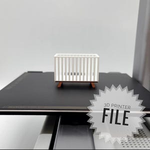 May include: A 3D printed model of a white cot with wooden legs. The model is on a black surface with a white starburst sticker that says "3D Printer File".