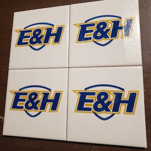May include: Set of four white ceramic coasters with a blue and gold logo that reads "E&H".