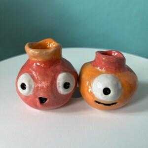 May include: Two small, handmade ceramic vases with glossy finishes. The left vase is pinkish-red with two large white eyes and a small black mouth. The right vase is orange with a single large white eye and a thin black line for a smiling mouth.