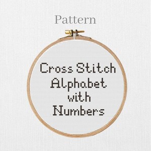 May include: A wooden embroidery hoop holds white fabric with a cross-stitch alphabet and numbers pattern. The text "Cross Stitch Alphabet with Numbers" is stitched in black thread. The word "Pattern" is above the hoop.