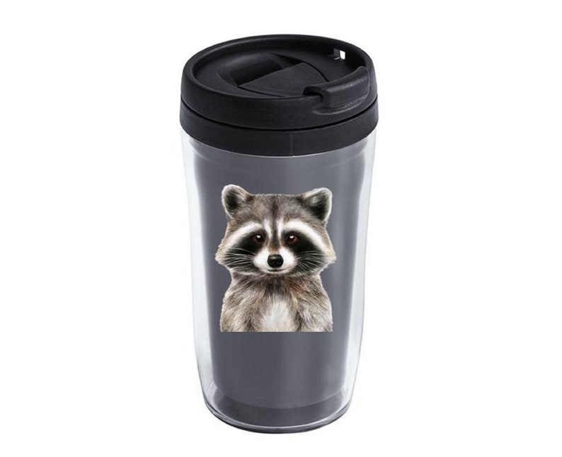 May include: A black and gray travel mug with a raccoon face design. The raccoon has black eyes, a black nose, and gray fur.