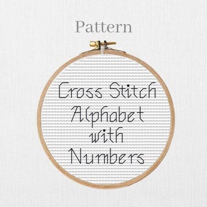 May include: A circular wooden embroidery hoop displaying a cross-stitch design. The design includes the text "Cross Stitch Alphabet with Numbers" in black thread on a grey and white background. The word "Pattern" is above the hoop.