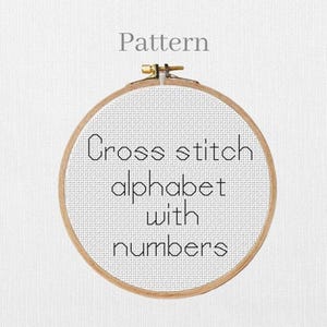 May include: A wooden embroidery hoop holding white fabric with a cross-stitch pattern. The text "Cross stitch alphabet with numbers" is displayed in black. The word "Pattern" is above the hoop.