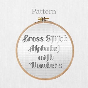 May include: A round wooden embroidery hoop containing white Aida fabric with black cross-stitched text that reads Cross Stitch Alphabet with Numbers. The word Pattern is printed in grey above the hoop on a white textured background.
