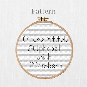 Op de afbeelding: Een kruissteekpatroon in een houten hoepel. De tekst luidt "Cross Stitch Alphabet with Numbers" in zwart, met het woord "Pattern" erboven. De stof heeft een grijs en wit gestreept patroon.