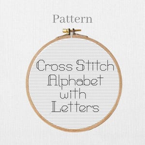 May include: A round wooden embroidery hoop containing white Aida fabric with black cross-stitched text that reads Pattern, Cross Stitch, Alphabet, with, and Letters. The hoop features a metal tension screw at the top center for fabric tightening.