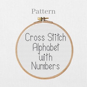 May include: A circular wooden embroidery hoop displaying a cross-stitch design. The design includes the text "Cross Stitch Alphabet with Numbers" in black. The word "Pattern" is above the hoop.