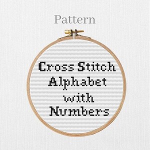 Può includere: Un modello a punto croce in un telaio di legno. Il testo "Cross Stitch Alphabet with Numbers" è ricamato con filo nero su sfondo bianco. La parola "Pattern" è sopra.